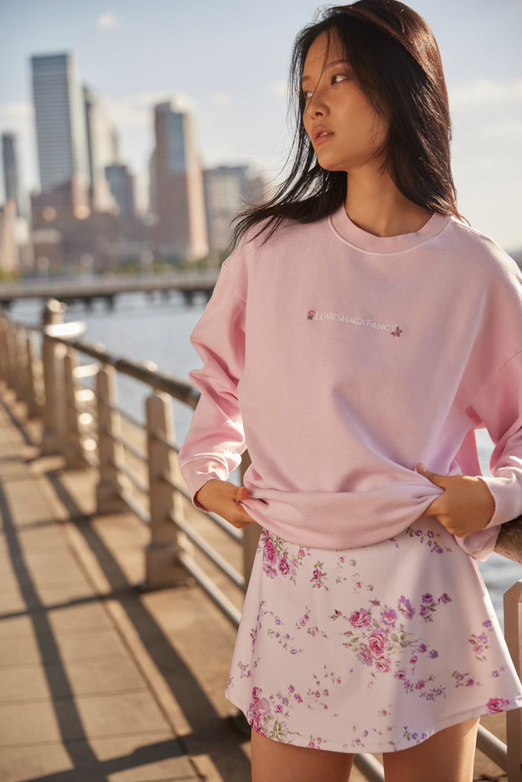 Pink crewneck sweatshirt with floral hem and chest logo, worn by model