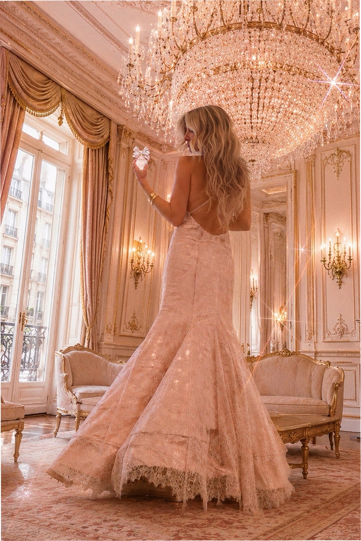 Woman in long pink tulle gown under chandeliers, wearing perfume bottle on display.