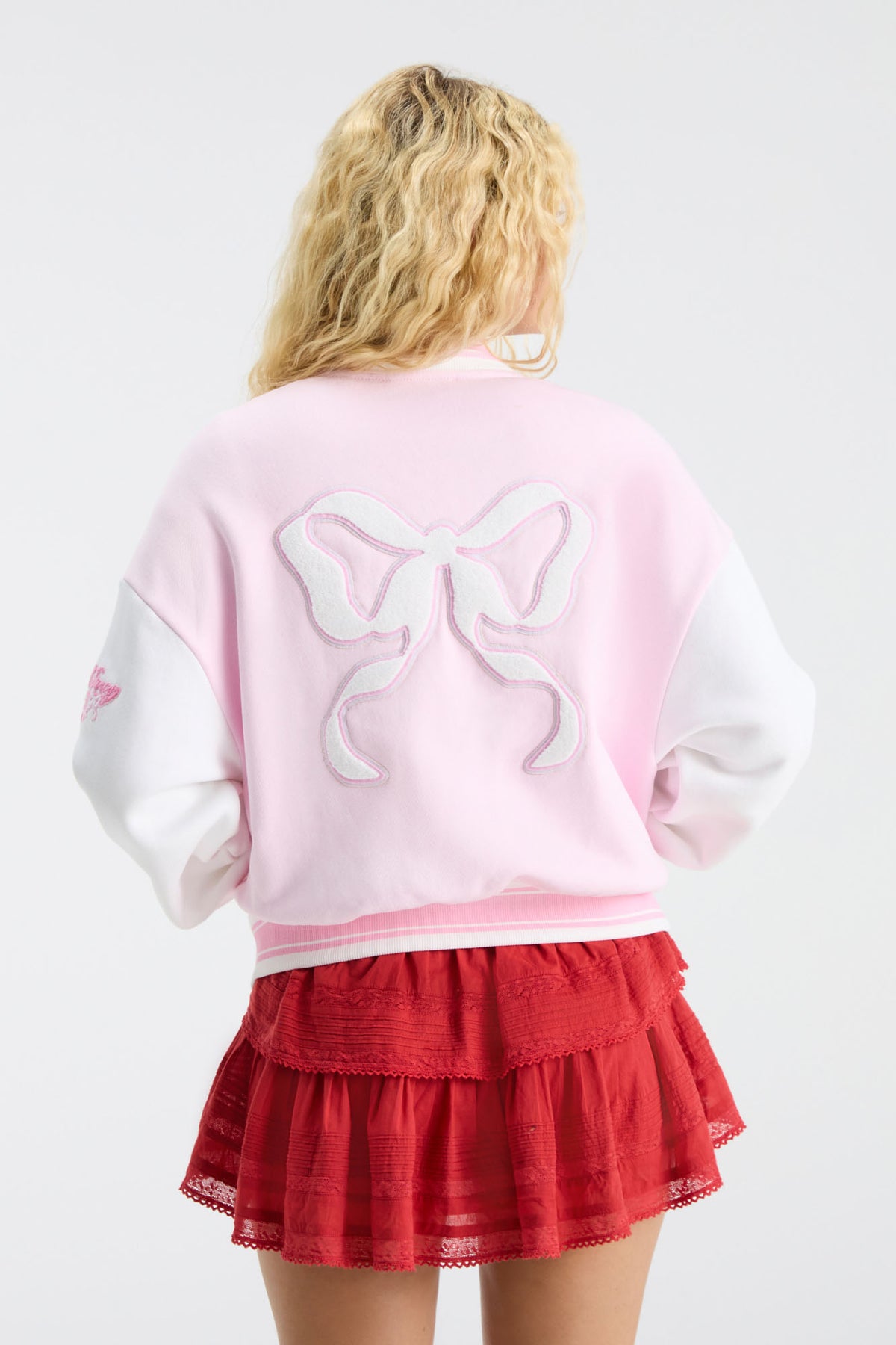 Back view of pink varsity jacket with bow emblem and striped rib trim over red skirt