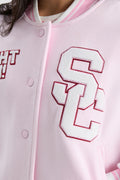 Light pink varsity jacket close-up with red-outlined letter patch and snap front.