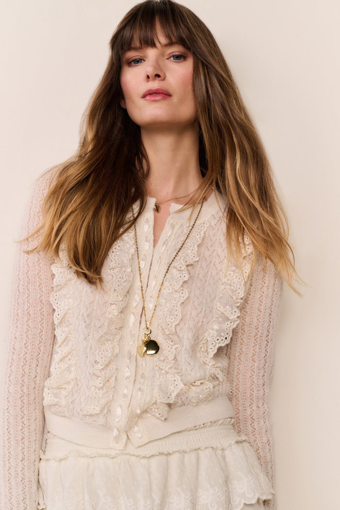 Woman wearing a knitted ruffle cream cardigan top with ruffle trim white pants on a plain background