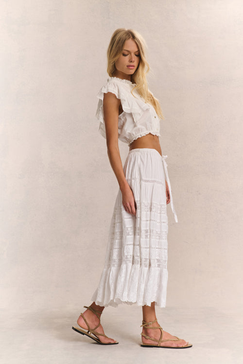 Woman wears white ruffle cropped top with midi white lace detail skirt against white background