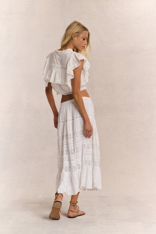 Woman wears white ruffle cropped top with midi white lace detail skirt against white background