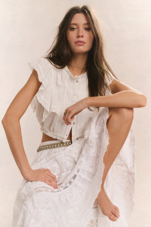 Woman wears white ruffle cropped top with midi white lace detail skirt against white background