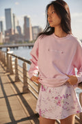 Pink crewneck sweatshirt with floral hem and chest logo, worn by model