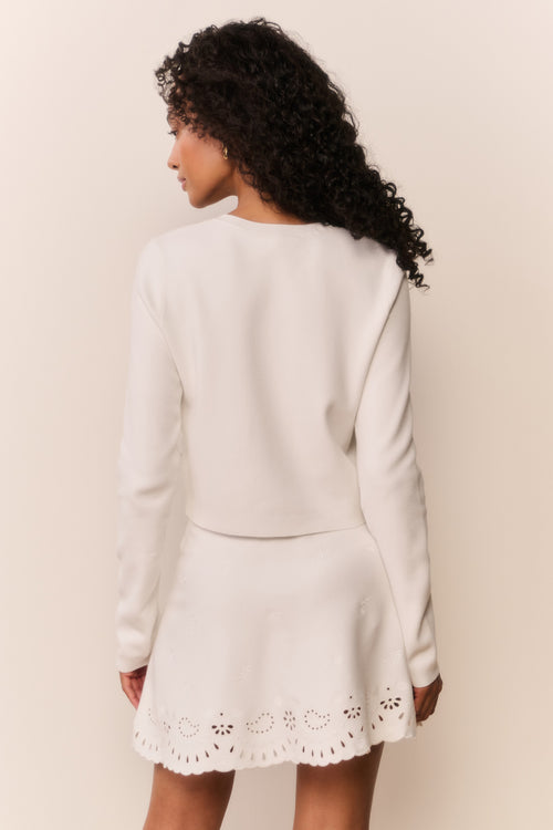 Woman wearing a white cardigan with intricate eyelet patterns and matching skirt on a plain background
