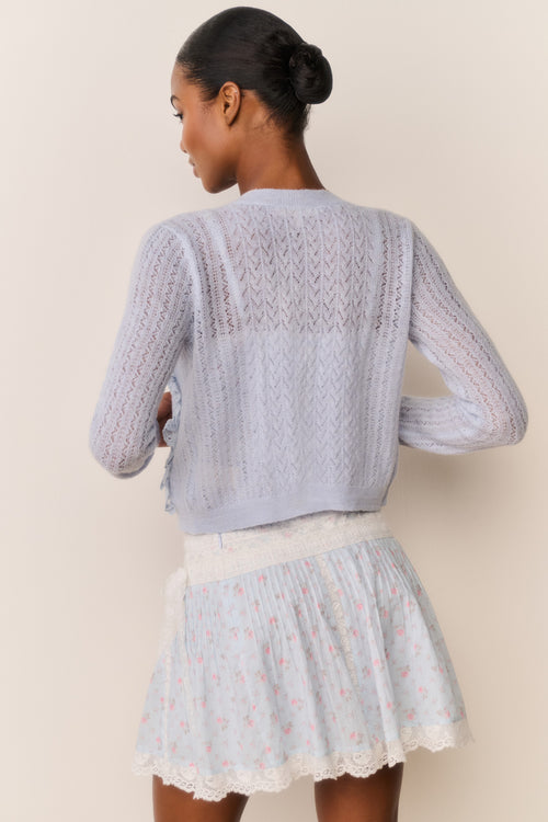 Woman wearing a light blue knitted cardigan over a blue skirt with floral patterns on a beige background