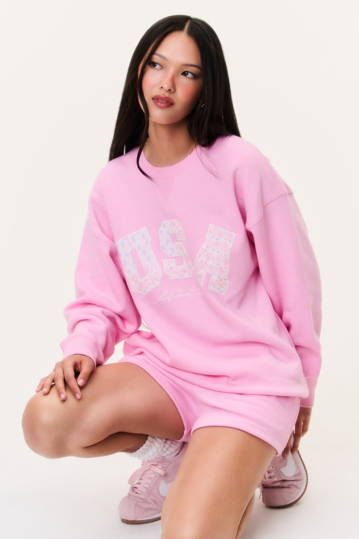 Pink fleece pullover on-model kneeling; large "USA" chest graphic and relaxed fit.
