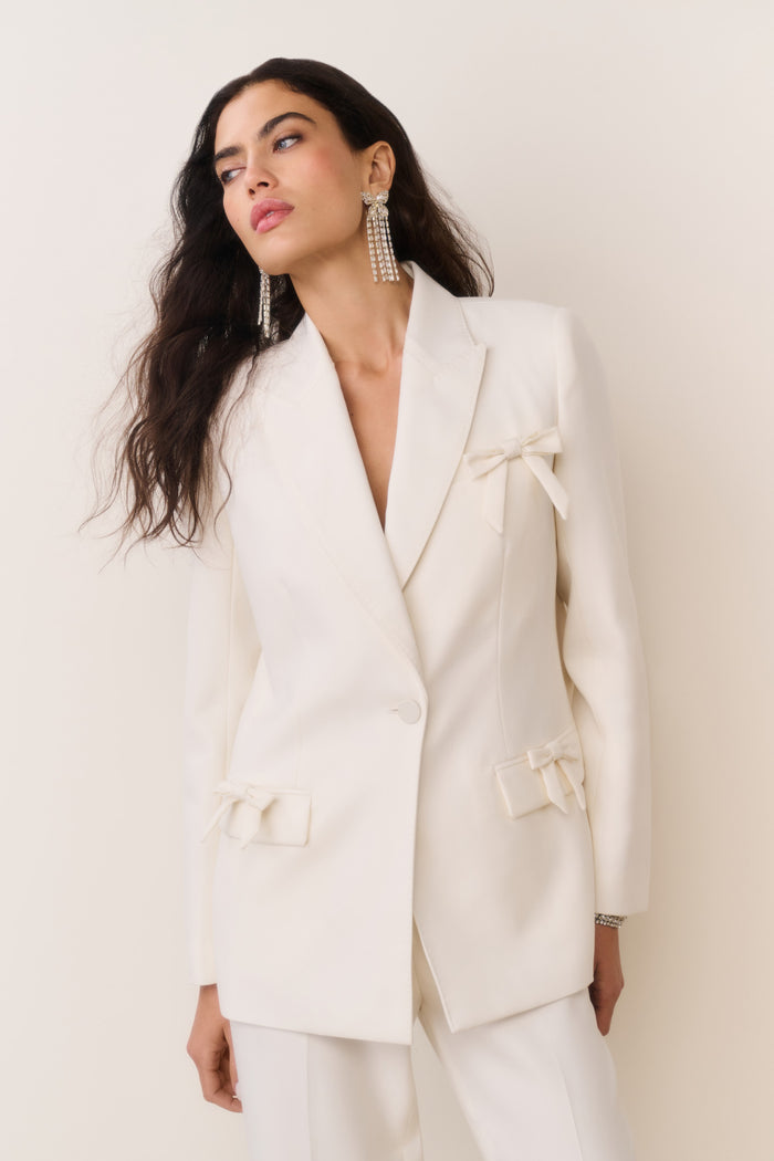 Cream wool-blend blazer with bow embellishments on lapel, cuffs, and flap pockets, close-up