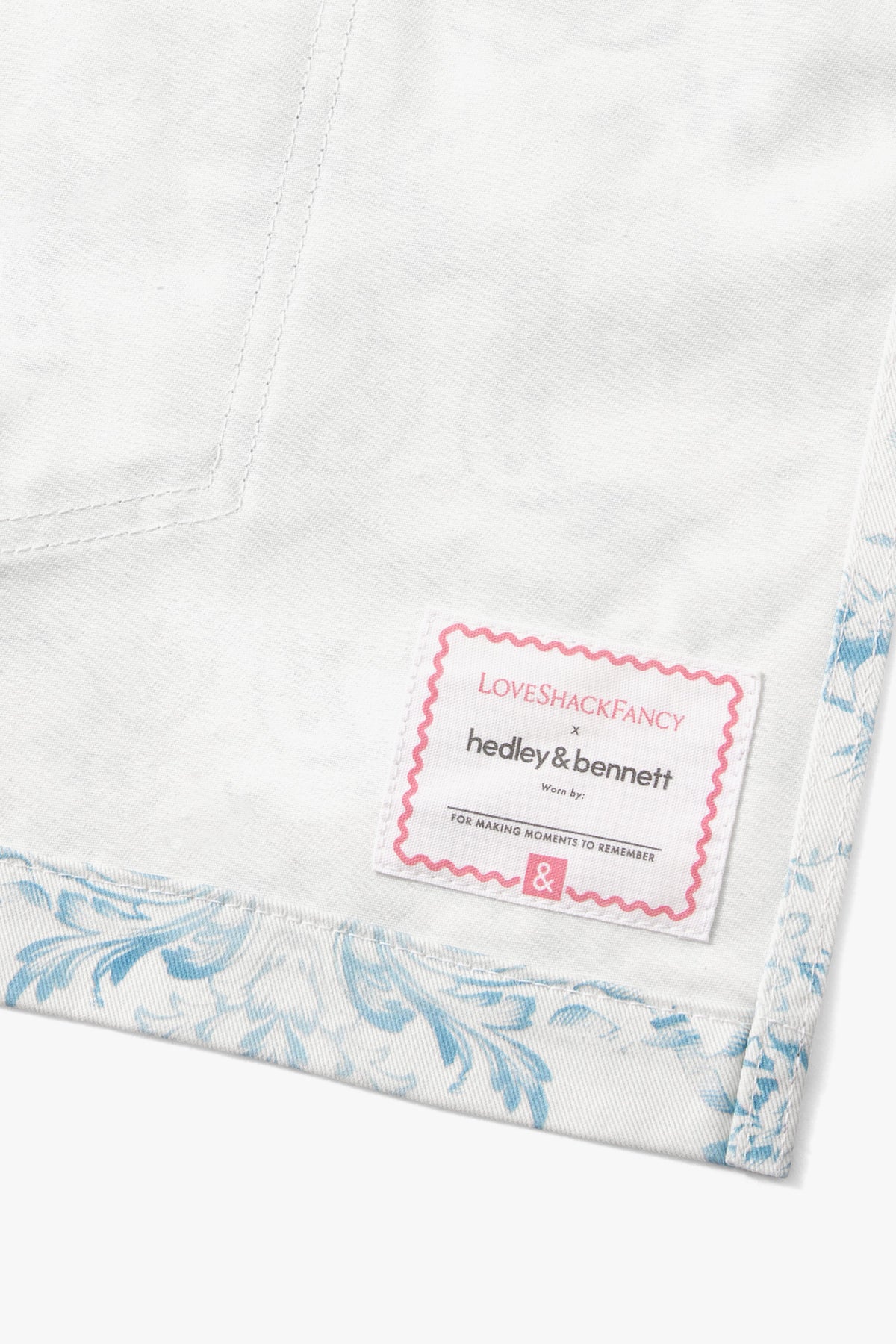 White apron with light blue print and pink brand label on hem